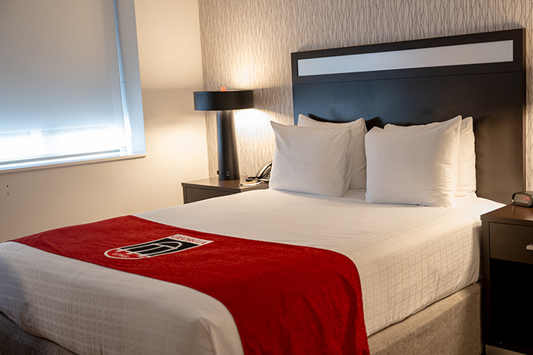 View of a hotel classic room with a bed and table lamp and the UGA shield on a red cloth at the foot of the bed.