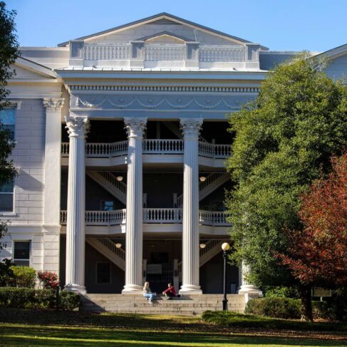 The front of the Holmes-Hunter Academic Building with students on the steps framed by North Campus trees turning red with colorful fall leaves.