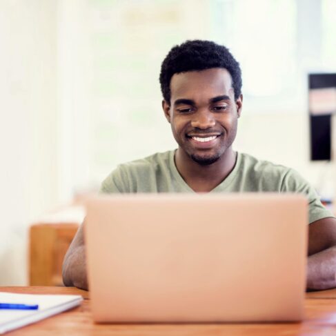 A man wearing a light green shirt is looking at his laptop computer with a notepad and a cup on each side in bright room