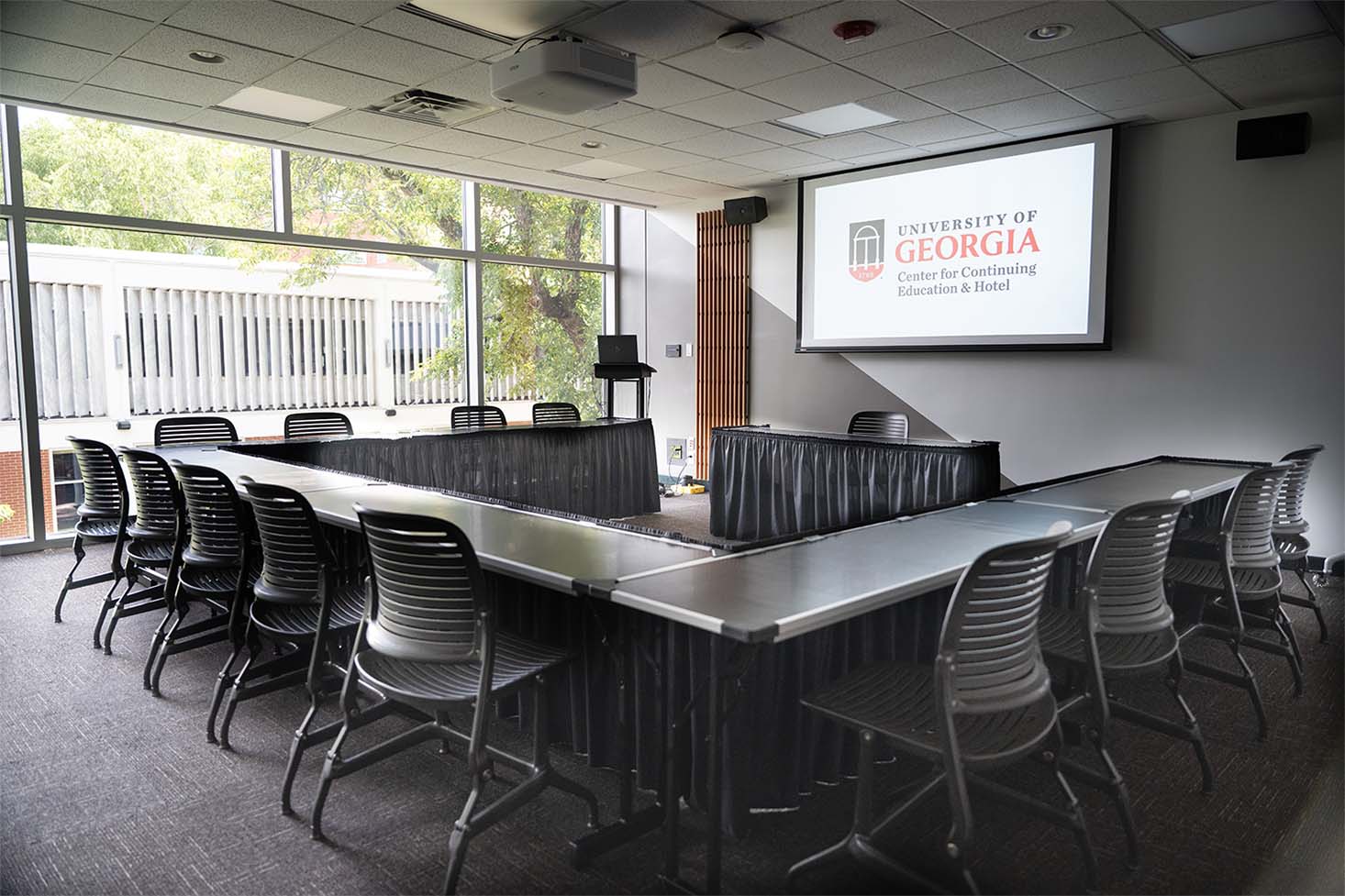 View inside a room that has a projection screen, white board, and tables in a U shape in front of a podium.