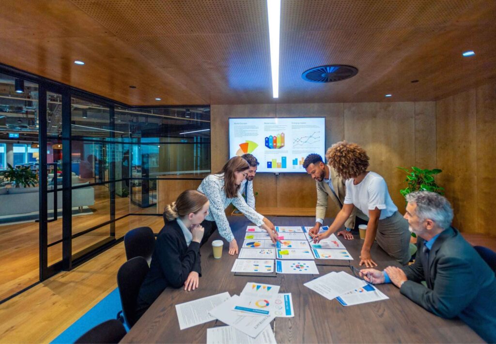 Paperwork and group of peoples hands on a board room table at a business presentation or seminar. The documents have financial or marketing figures, graphs and charts on them. People are pointing at the data. There is a T.V. with financial data in the background