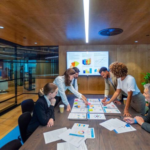 Paperwork and group of peoples hands on a board room table at a business presentation or seminar. The documents have financial or marketing figures, graphs and charts on them. People are pointing at the data. There is a T.V. with financial data in the background