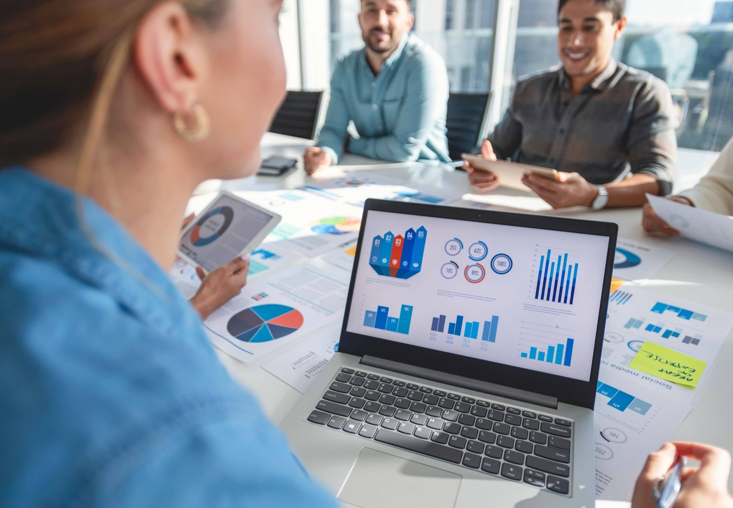 Group of people working with paperwork on a board room table at a business presentation or seminar. The documents have financial or marketing figures, graphs and charts on them. There is a laptop with graph and chart infographic data