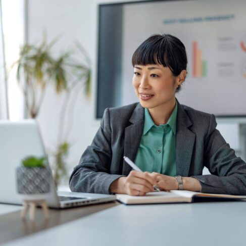 A businesswoman in a corporate suit takes notes as she participates in a meeting, with a laptop and data charts in the background for visual reference.