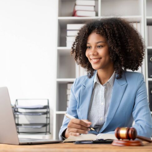 A female professional sits at a desk in an office, holding a pen and reviewing documents. An open laptop is on the left, and a gavel rests on papers in front of them. Shelves with books and binders are visible in the background.