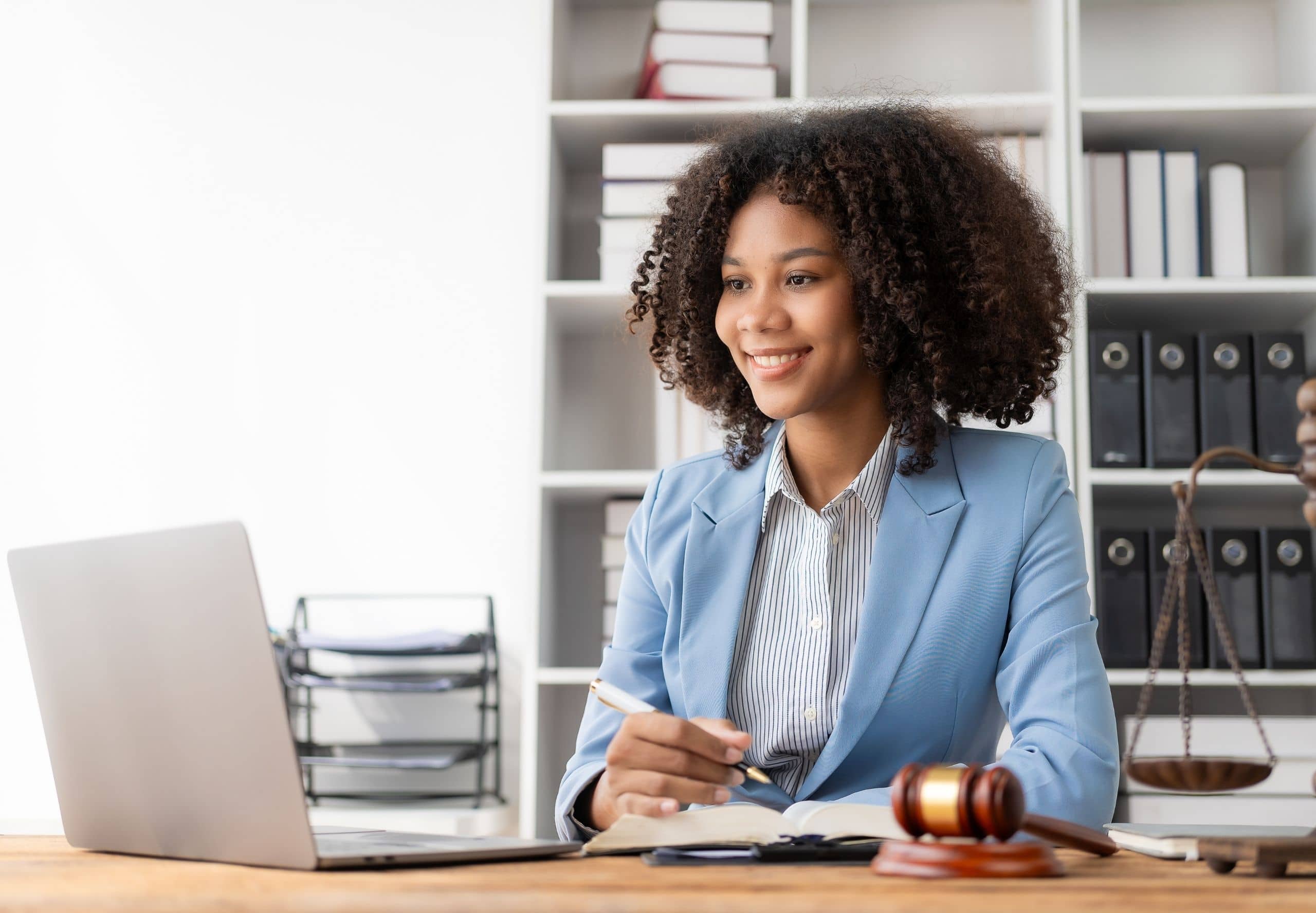 A female professional sits at a desk in an office, holding a pen and reviewing documents. An open laptop is on the left, and a gavel rests on papers in front of them. Shelves with books and binders are visible in the background.