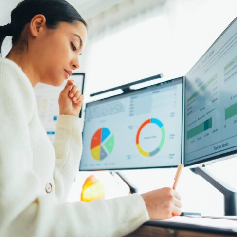 Woman working at her desk writing notes as she goes over graphics, charts and data tables displayed on her computer screens
