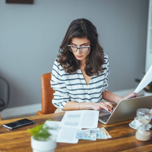 A woman sitting at a wooden table, holding papers in one hand and typing on a laptop with the other.