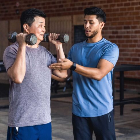 An adult male personal trainer helps an active senior man use hand weights while working out in a cross training gym.