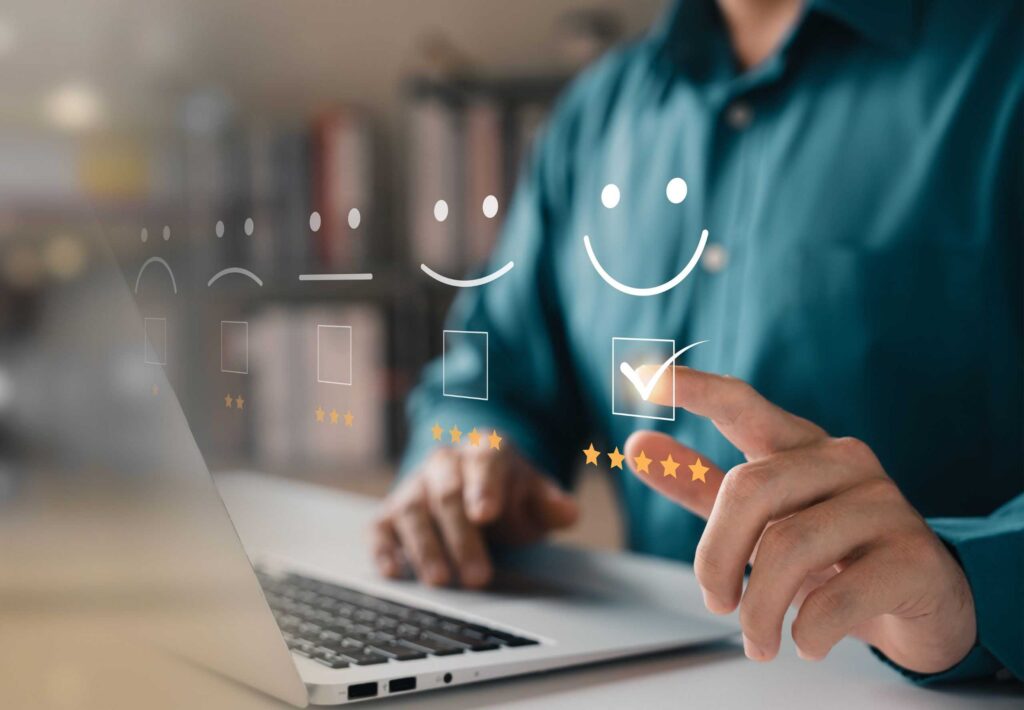 A person uses a laptop and points at a virtual interface displaying five satisfaction levels represented by smiley faces, ranging from sad to very happy. The person selects the happiest face, which is accompanied by five stars."
