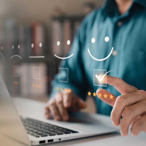 A person uses a laptop and points at a virtual interface displaying five satisfaction levels represented by smiley faces, ranging from sad to very happy. The person selects the happiest face, which is accompanied by five stars."