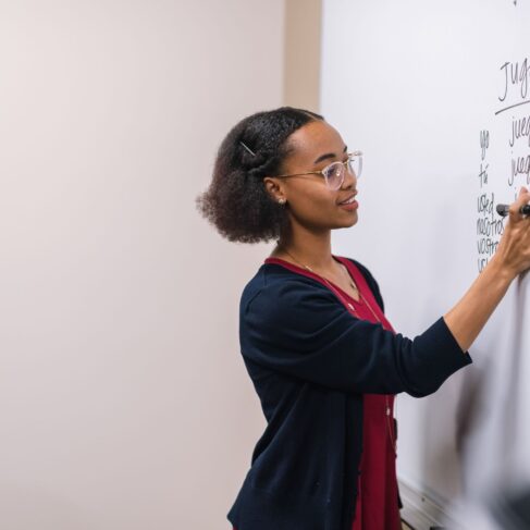 A female professor stands at a large whiteboard and writes down Spanish verbs to conjugate.