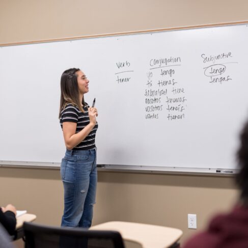 Teacher teaching online class with a white board in the back with Spanish verbs written on it.