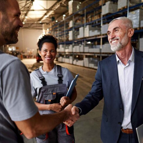 A happy manager and warehouse employee shaking hands next to female colleague in distribution warehouse.