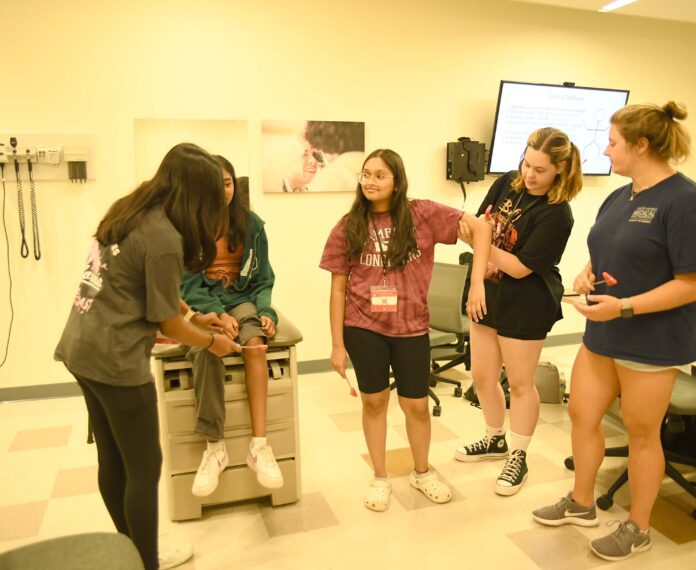 A group of students practicing medical rounds use a reflex hammer on a practice patient