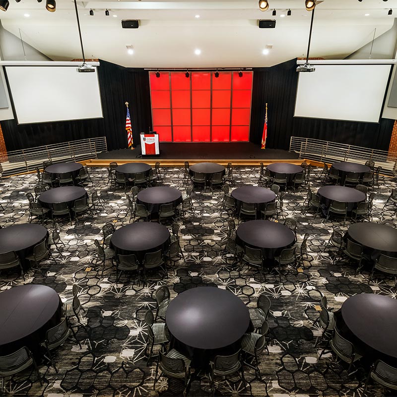 View of Mahler Hall with round tables and two projection screens and a podium with flags on each side of the stage.