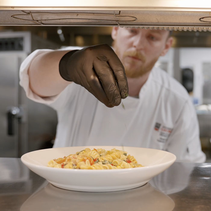 Dustin Ellis sprinkling parsley on a pasta dish in the kitchen.