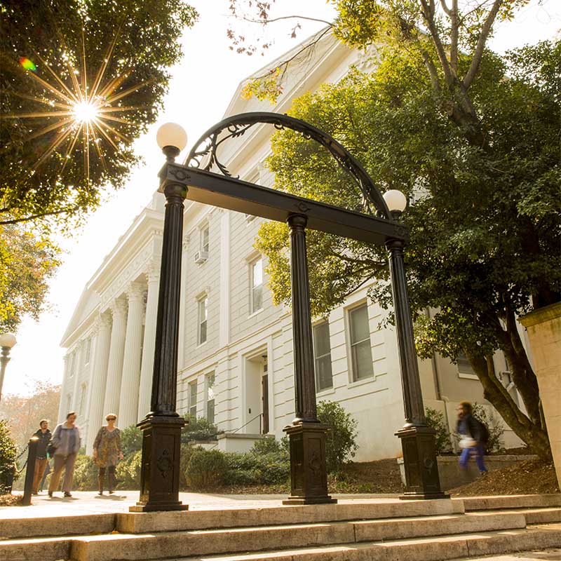 View of the University of Georgia Arch on the campus with a white building in the background and people walking.