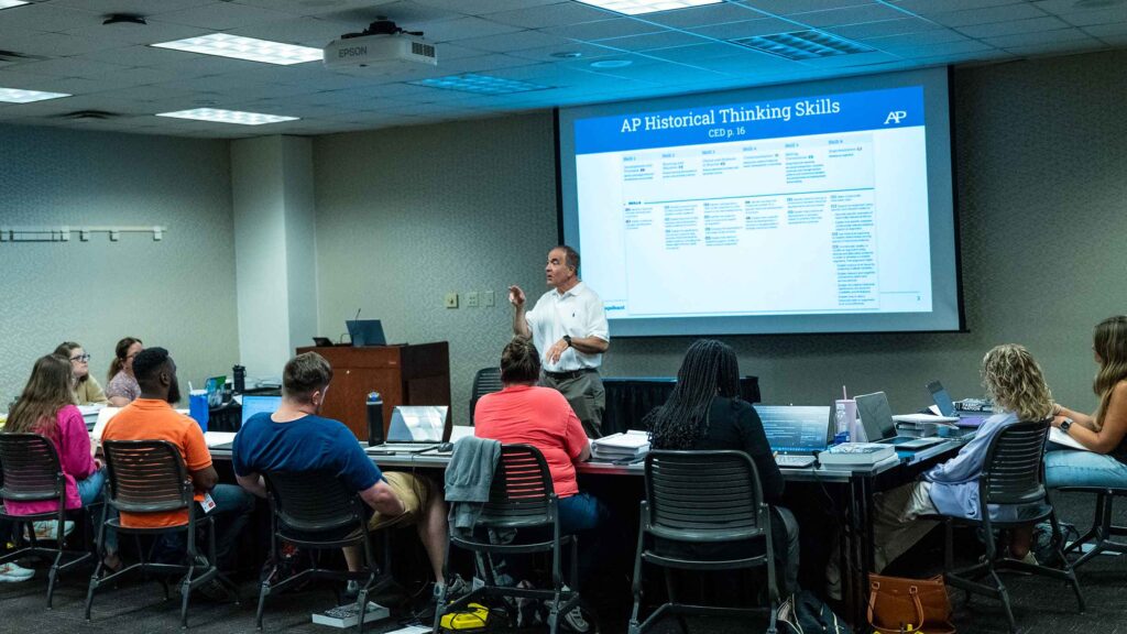 An instructor teaching about AP Historical Thinking Skills which is displayed on the screen to a group of students in a classroom.