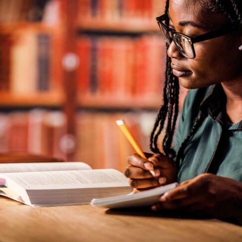 Diligent young woman sitting at table in a library, listening to music via blue-tooth headphones, reading a book and taking notes while studying for exam.