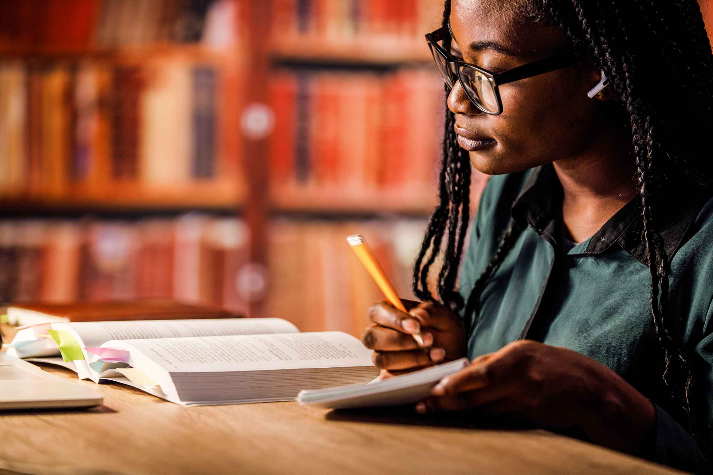 Diligent young woman sitting at table in a library, listening to music via blue-tooth headphones, reading a book and taking notes while studying for exam.