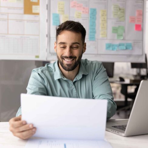 Positive satisfied business professional man in casual reading paper report at workplace with laptop, checking legal financial documents, letter with good news, smiling