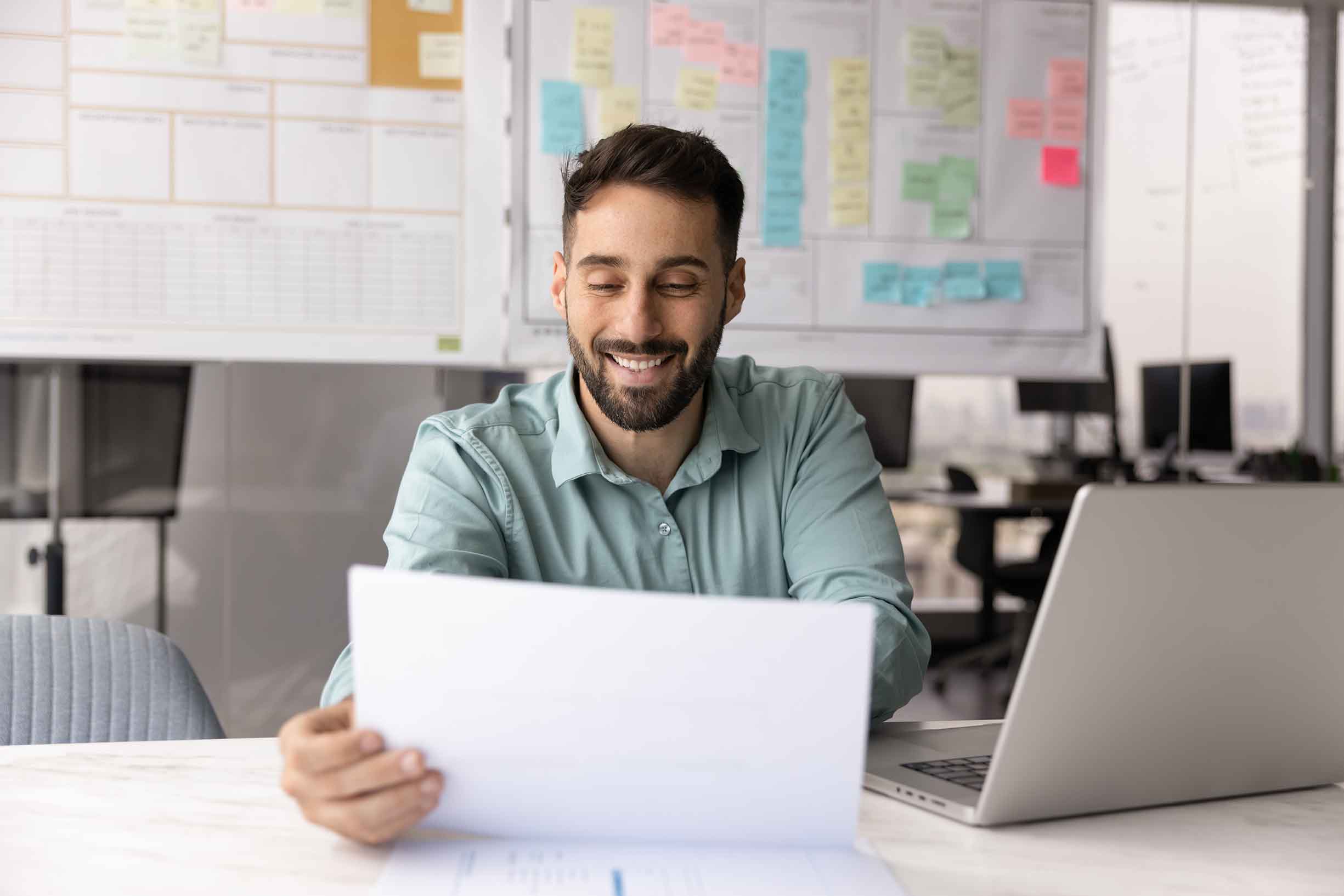Positive satisfied business professional man in casual reading paper report at workplace with laptop, checking legal financial documents, letter with good news, smiling