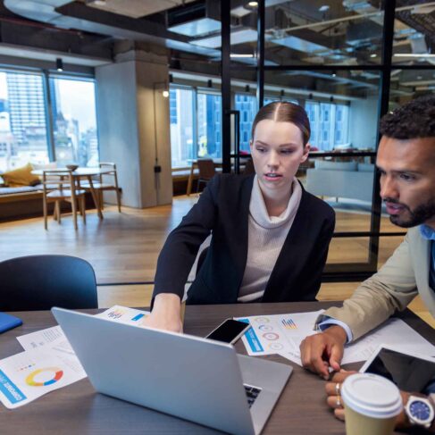 Two business people meeting in the office. There is a laptop on the table with paperwork showing financial graphs and figures. There is also coffee and other technology on the desk