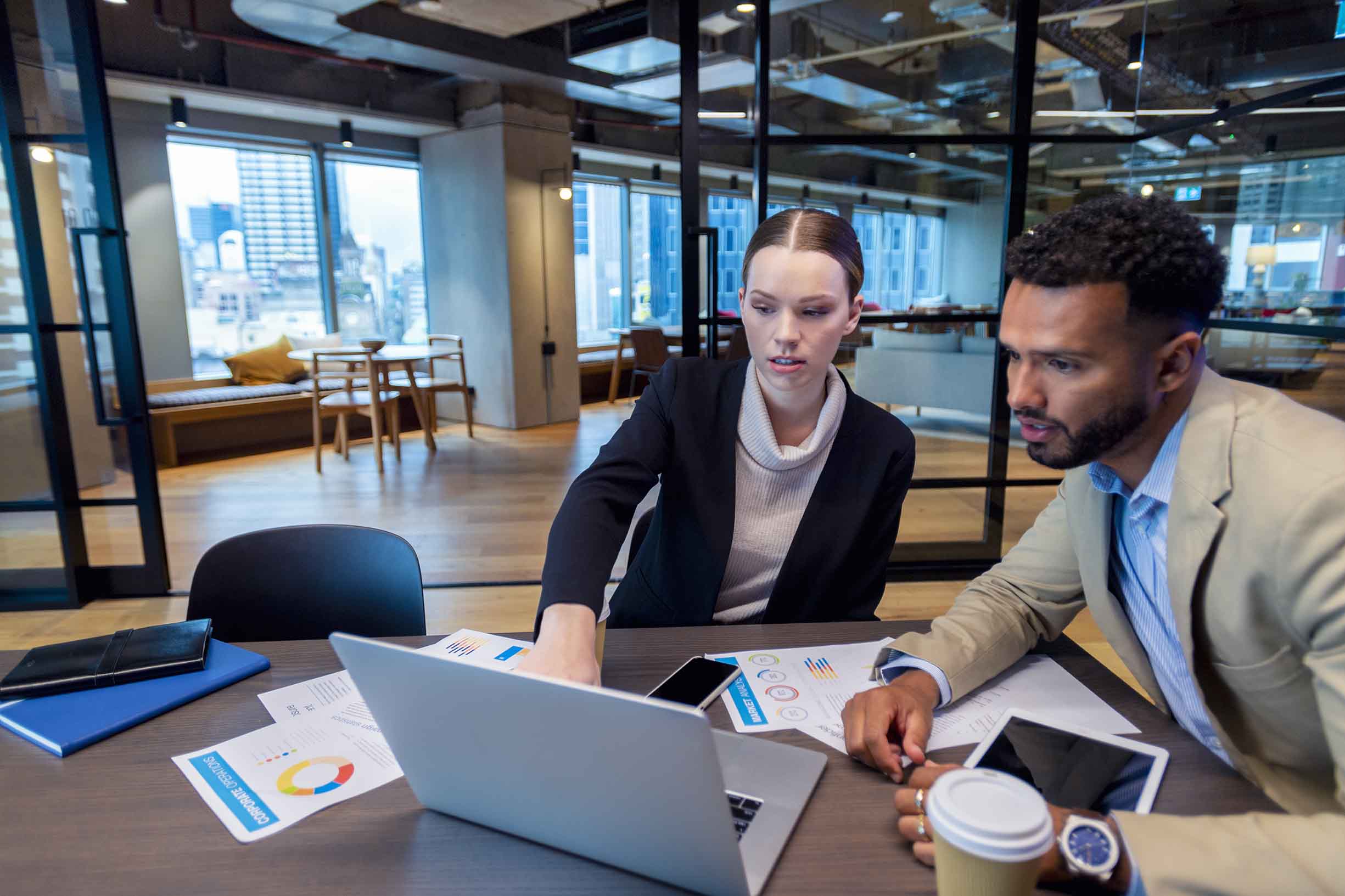 Two business people meeting in the office. There is a laptop on the table with paperwork showing financial graphs and figures. There is also coffee and other technology on the desk