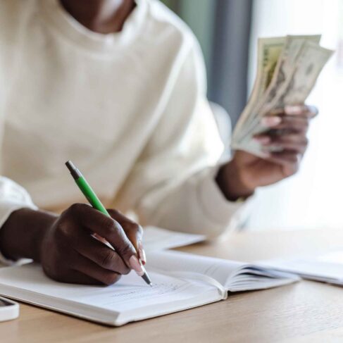 Cropped shot of a woman holding money while sitting at the table and writing expenses