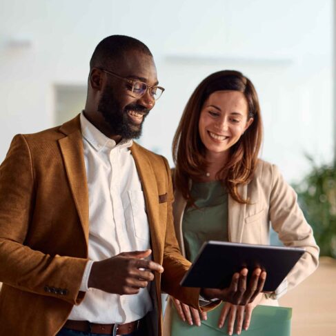 Two happy business colleagues engaging with a digital tablet and discussing various projects while standing in a bright, modern office environment, showcasing teamwork and collaboration