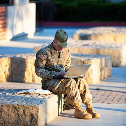 US soldier studying on laptop in front of campus.