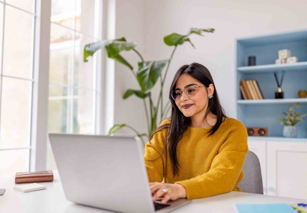 A concentrated woman in a mustard sweater works intently on her laptop in a serene, plant-filled home office, embodying dedication and tranquility in her tasks.