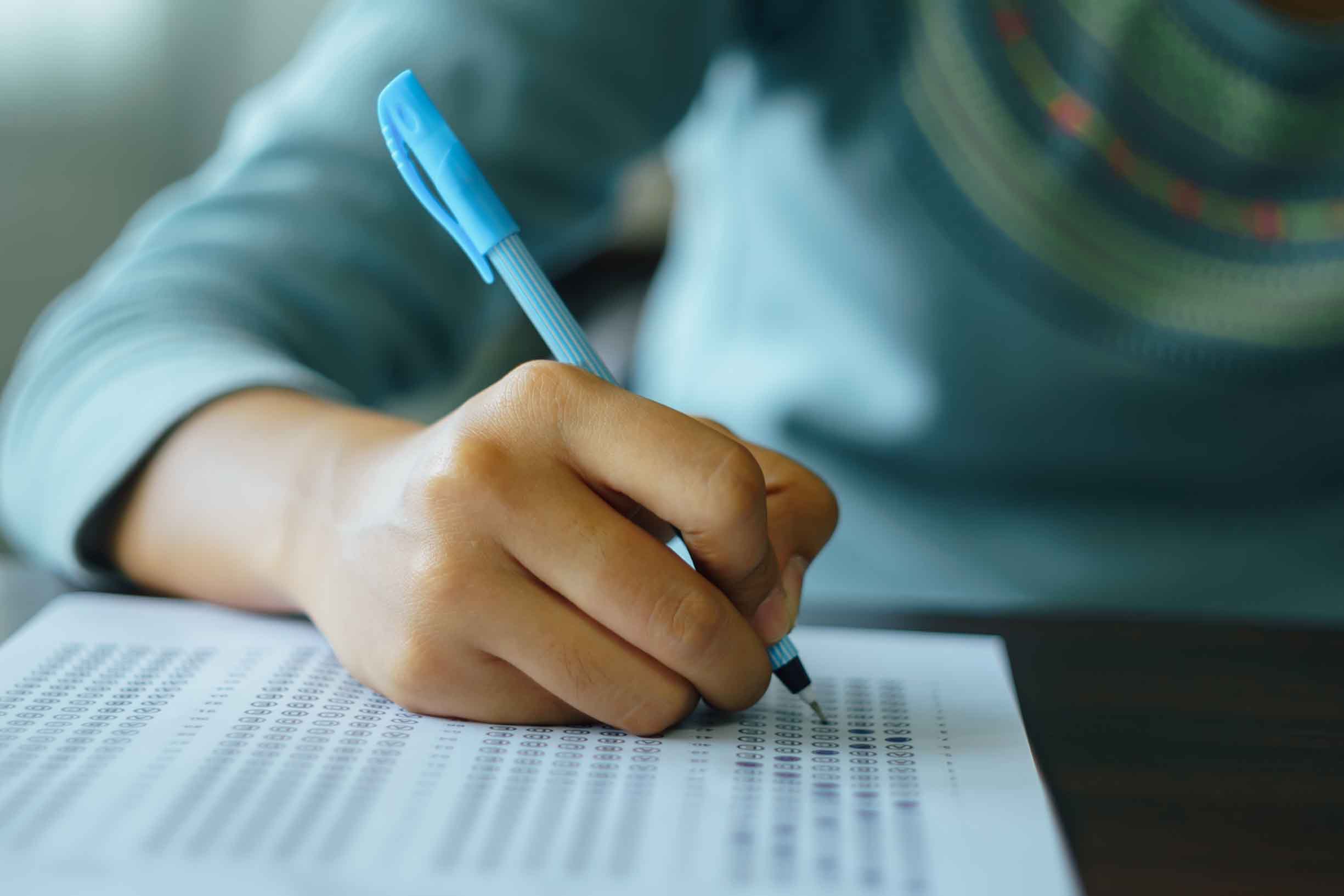 Close up of high school student holding a pen writing on answer sheet paper in the examination room.