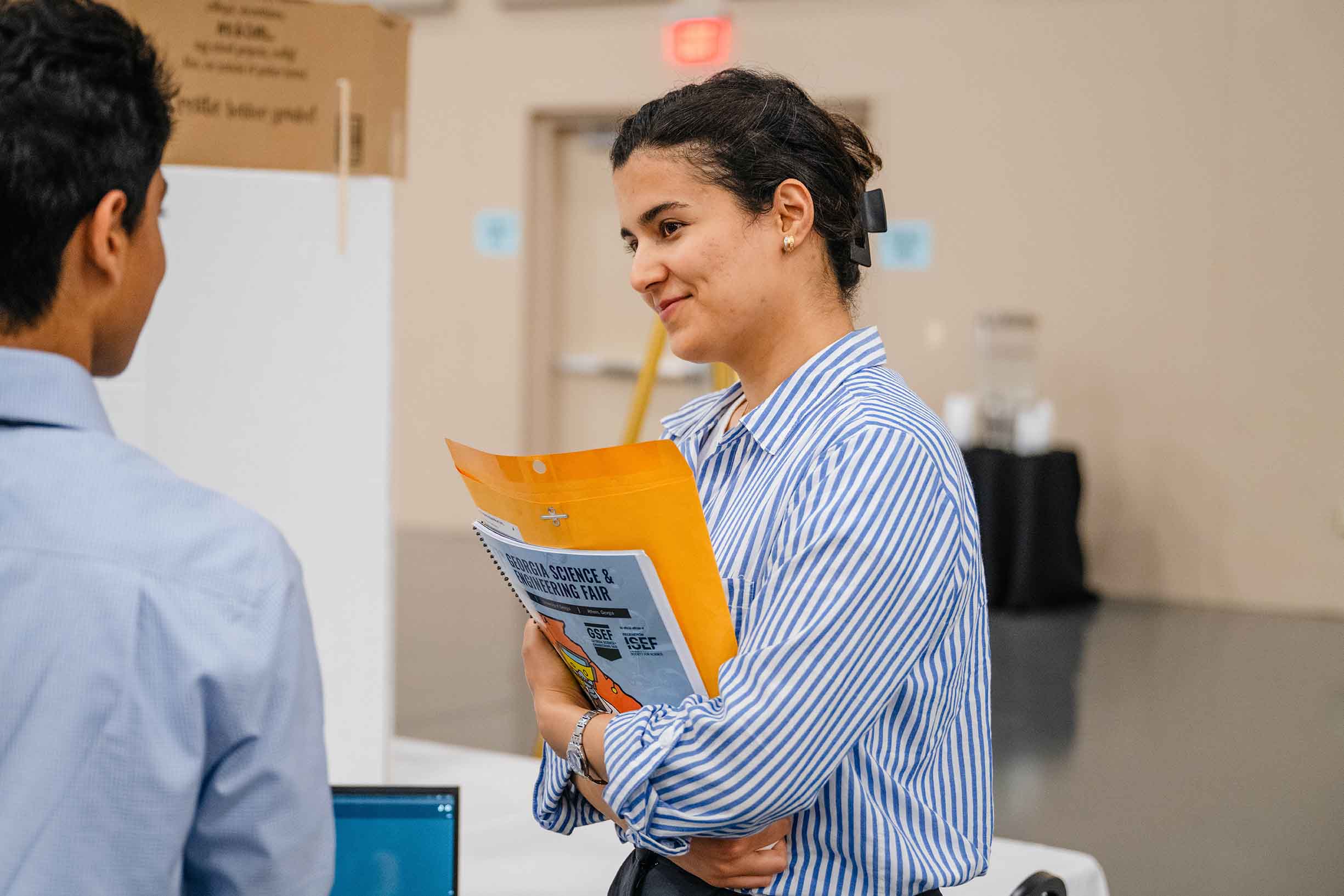 A female student holding her science fair information, listening to a student explaining his project