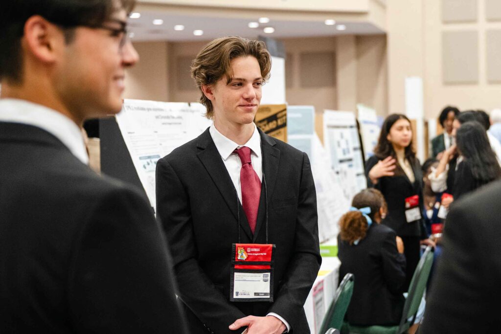 A science fair student dressed in a black suit with red tie standing in front of his project.