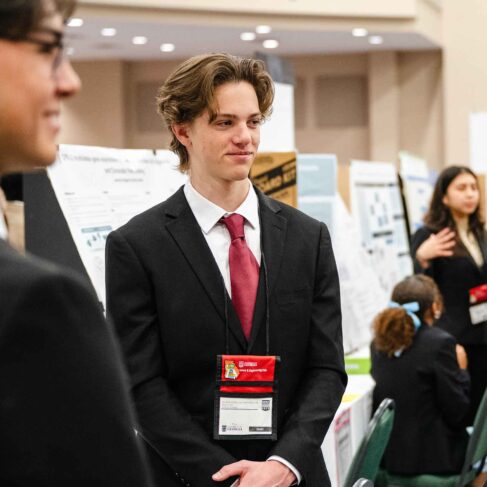 A science fair student dressed in a black suit with red tie standing in front of his project.