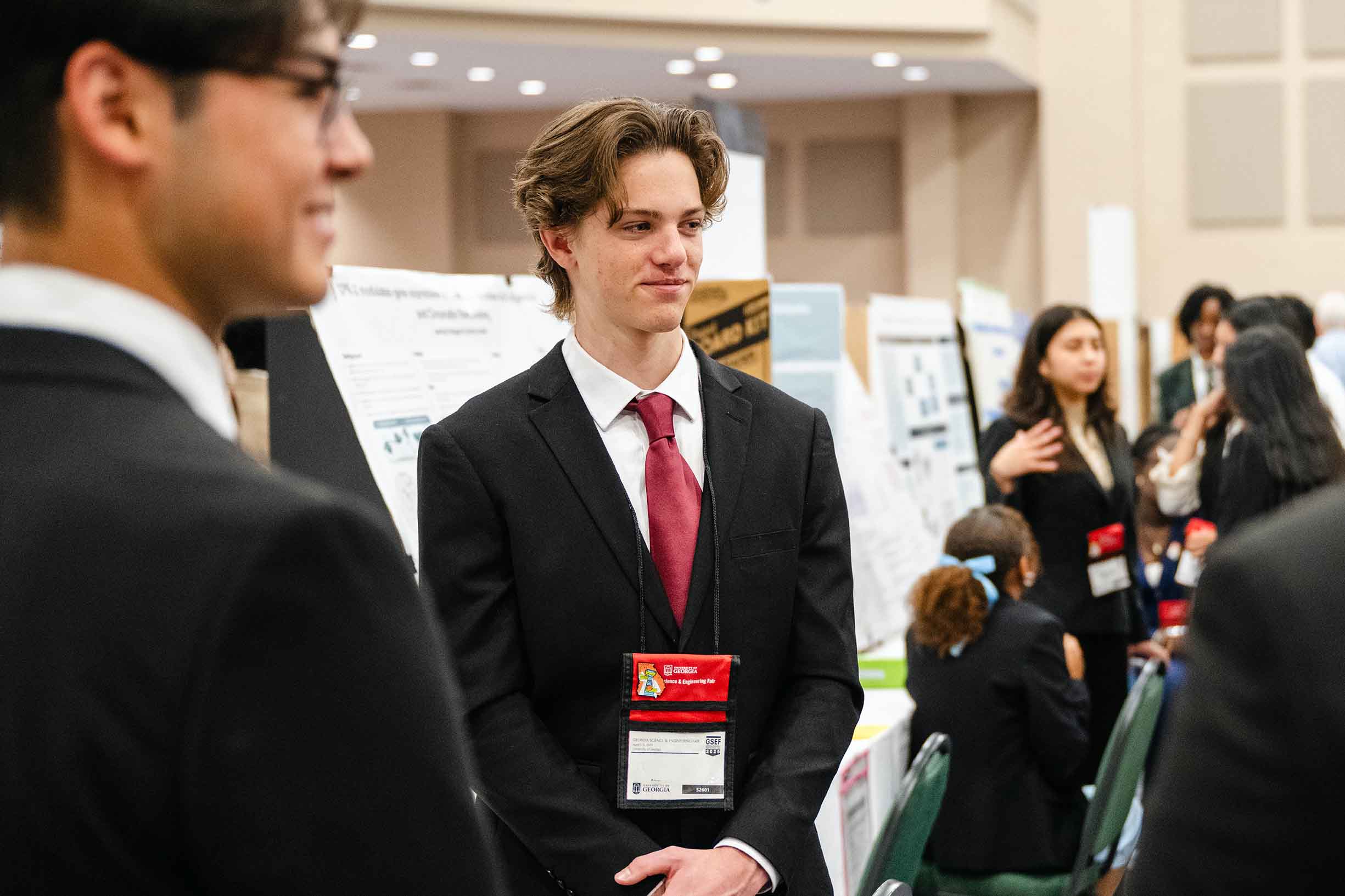 A science fair student dressed in a black suit with red tie standing in front of his project.