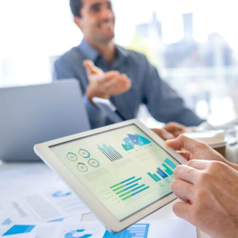 Group of men working with Paperwork on a board room table at a business meeting or seminar. The documents have financial or marketing figures, graphs and charts on them. They are all using laptops and digital tablets