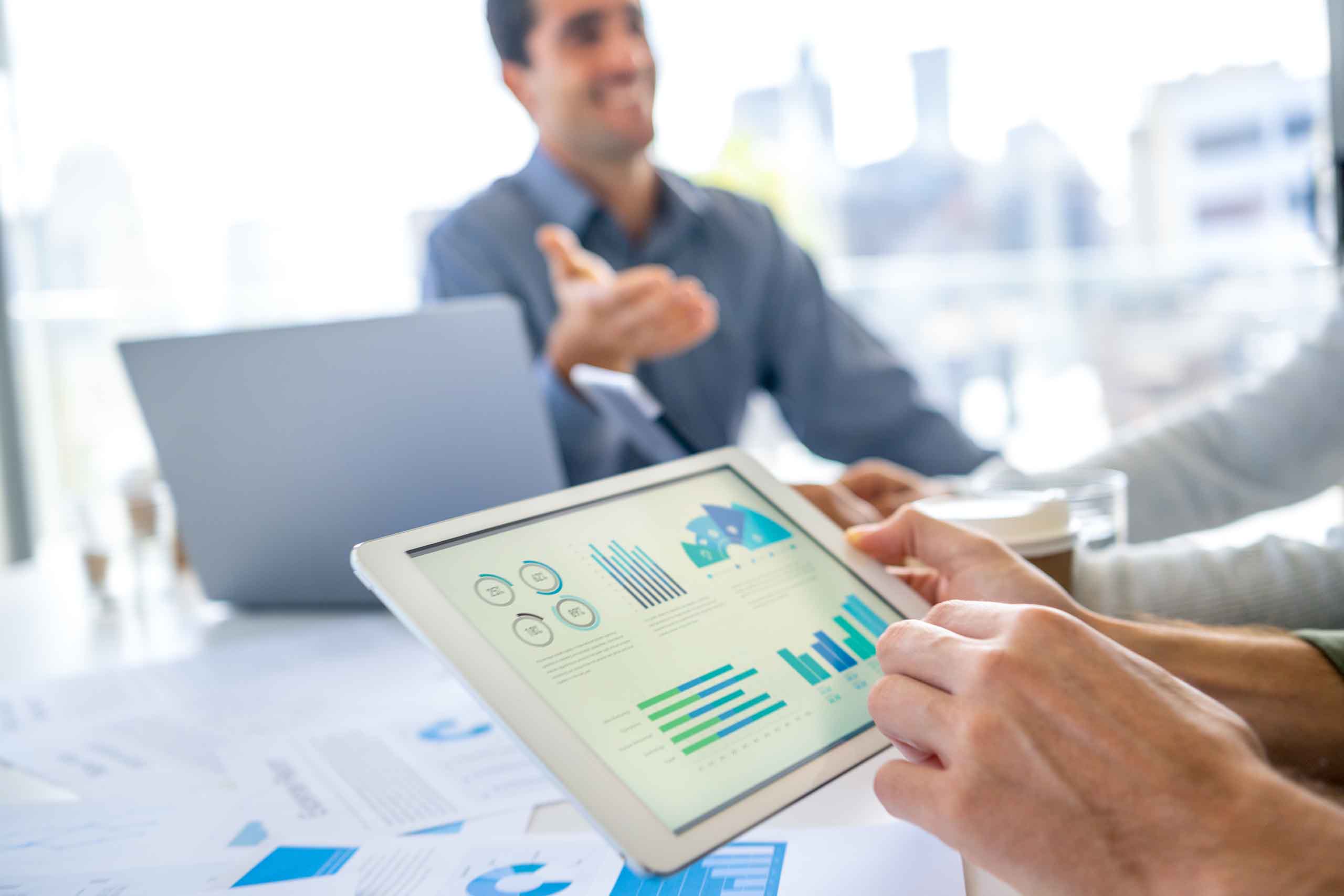 Group of men working with Paperwork on a board room table at a business meeting or seminar. The documents have financial or marketing figures, graphs and charts on them. They are all using laptops and digital tablets