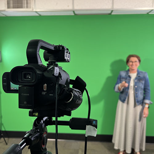 A female standing in front of a camera with a green screen behind her
