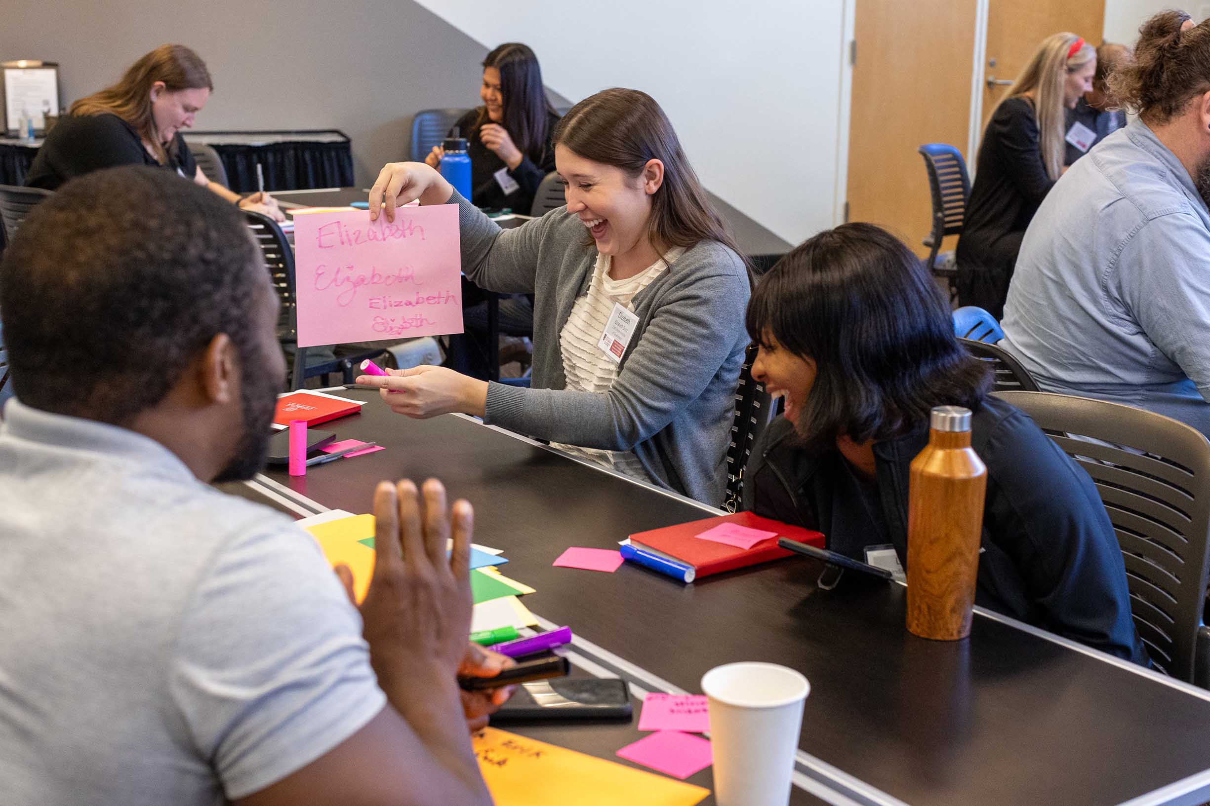 A PSO attendee holding a sheet of pink paper that says "Elizabeth and showing it to others sitting at a table