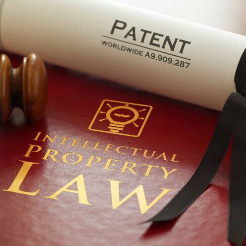 Red leather Intellectual Property Law book with with gold embossed type and iconic light bulb, ideas icon logo, with a judges gavel and a generic A4 Patent document. Note patent number is fictitious.