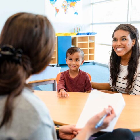 Sitting at a small table in the kindergarten classroom, the mid adult mother smiles as she discusses her young son's progress with the unrecognizable female teacher.