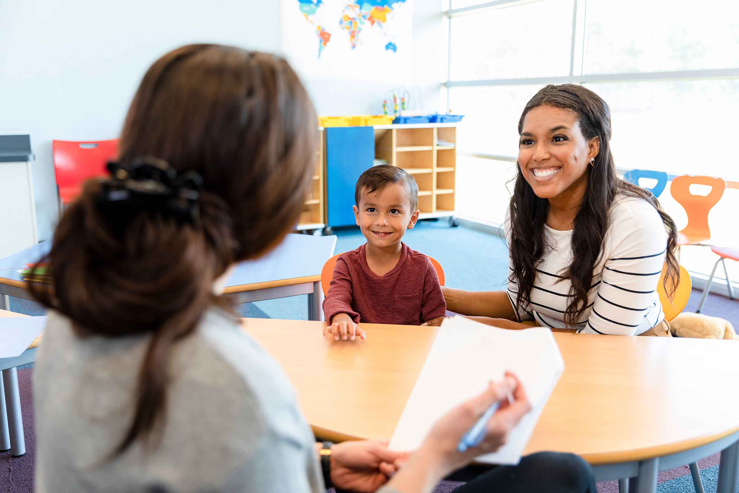 Sitting at a small table in the kindergarten classroom, the mid adult mother smiles as she discusses her young son's progress with the unrecognizable female teacher.