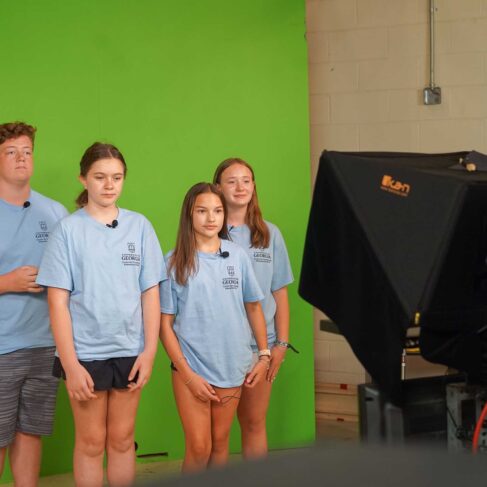 Four students wearing light blue shirts are standing in front of a green screen and camera to make a broadcast.