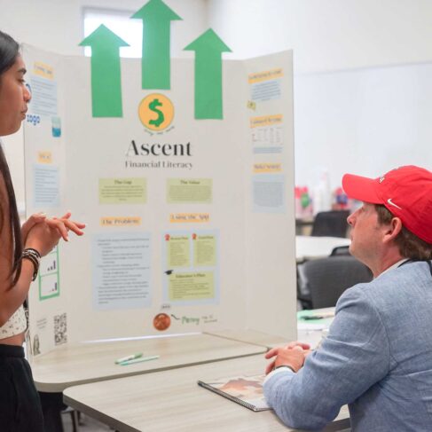 Student showing and explaining her financial plan board to her instructor in a classroom with tables