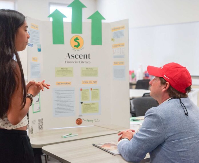 Student showing and explaining her financial plan board to her instructor in a classroom with tables