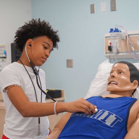 Student, wearing a white shirt, is listening to a patient's breathing in a mock hospital room.