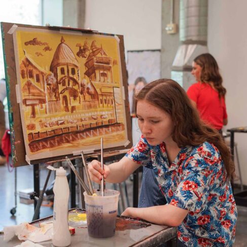 Female student painting a picture of building using yellows and brown colors in a classroom with other students in the background.